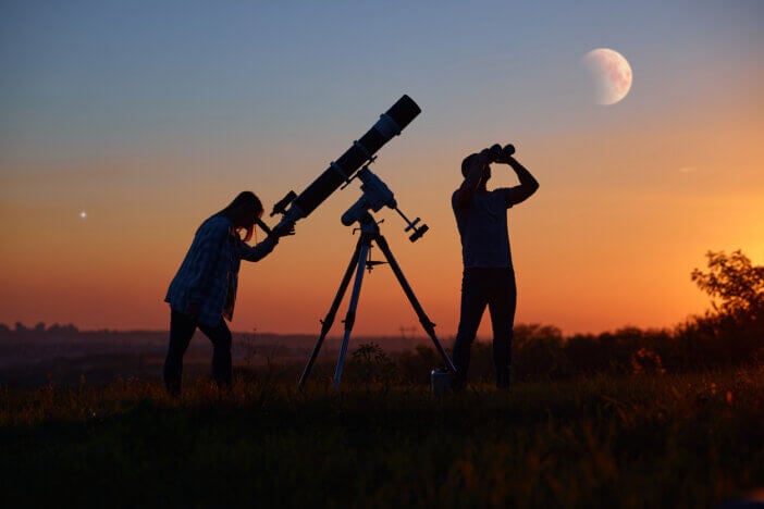 Couple stargazing together with a astronomical telescope, looking at planets, stars, lunar eclipse and meteor shower.